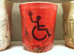 A ceramic mug with the "disabled" signage of a stick figure wheelchair user throwing a bomb.