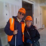 Dmitri Belser and Arlene Mayerson wearing hardhats in front of unpainted drywall.